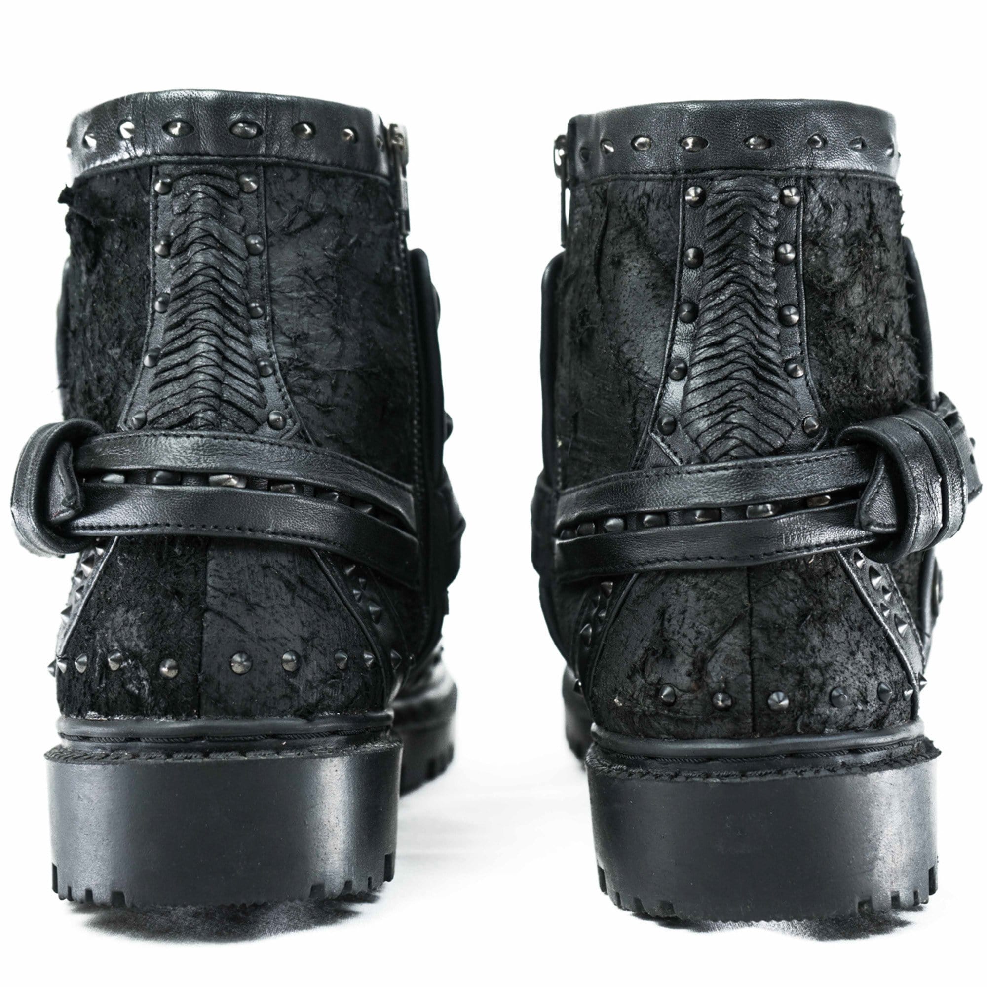 Introducing the Kaya Movement MAGMA Wasteland Studded Leather Ankle Combat Boots, a pair of rugged black punk rock boots showcasing intricate stitching, textured leather, and metallic studs. From the back, you'll notice decorative straps with buckles and thick, sturdy soles that beautifully complement their distressed finish for a stylishly worn look.